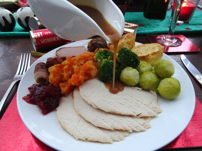 Photo showing some thick homemade gravy being poured over the roast turkey and vegetables of a Christmas dinner, from a white china gravy jug. The vegetables include Brussels sprouts and a mashed mixture of boiled carrots and swede.
